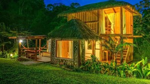 Una casa de madera de dos pisos con techo de paja, rodeada de abundante vegetación.