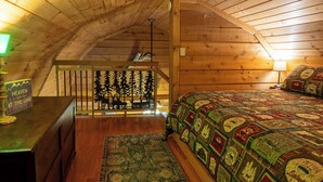 A cozy attic bedroom with wooden walls, a bed with a quilt, a desk with a lamp, and a decorative sign.
