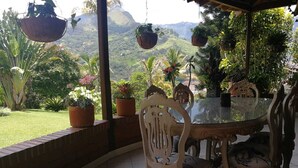 An outdoor dining area with a glass table and white chairs, surrounded by potted plants and a view of a mountainous landscape. - Hotel campestre (Copacabana)