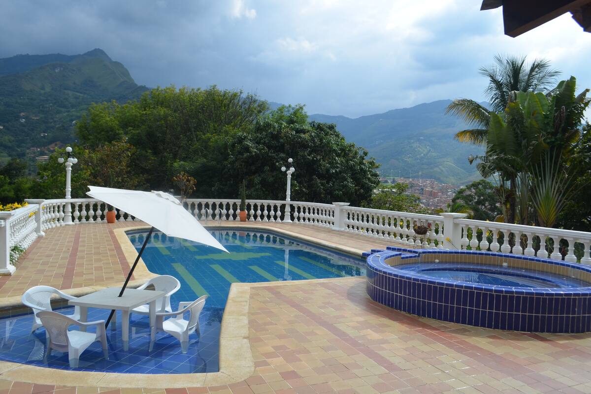 A swimming pool with a view of mountains, a hot tub, and a covered seating area with a table and chairs.