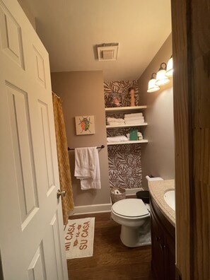 A bathroom with a toilet, sink, and shelves with towels. There is a sign on the floor and a painting on the wall. - Cozy, cabin in the woods perfect for hanging out. (Savannah)