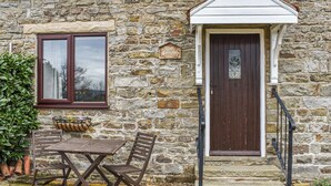 Une petite maison en pierre avec une table et des chaises en bois à l’extérieur, une fenêtre et une porte surmontée d’un petit porche.
