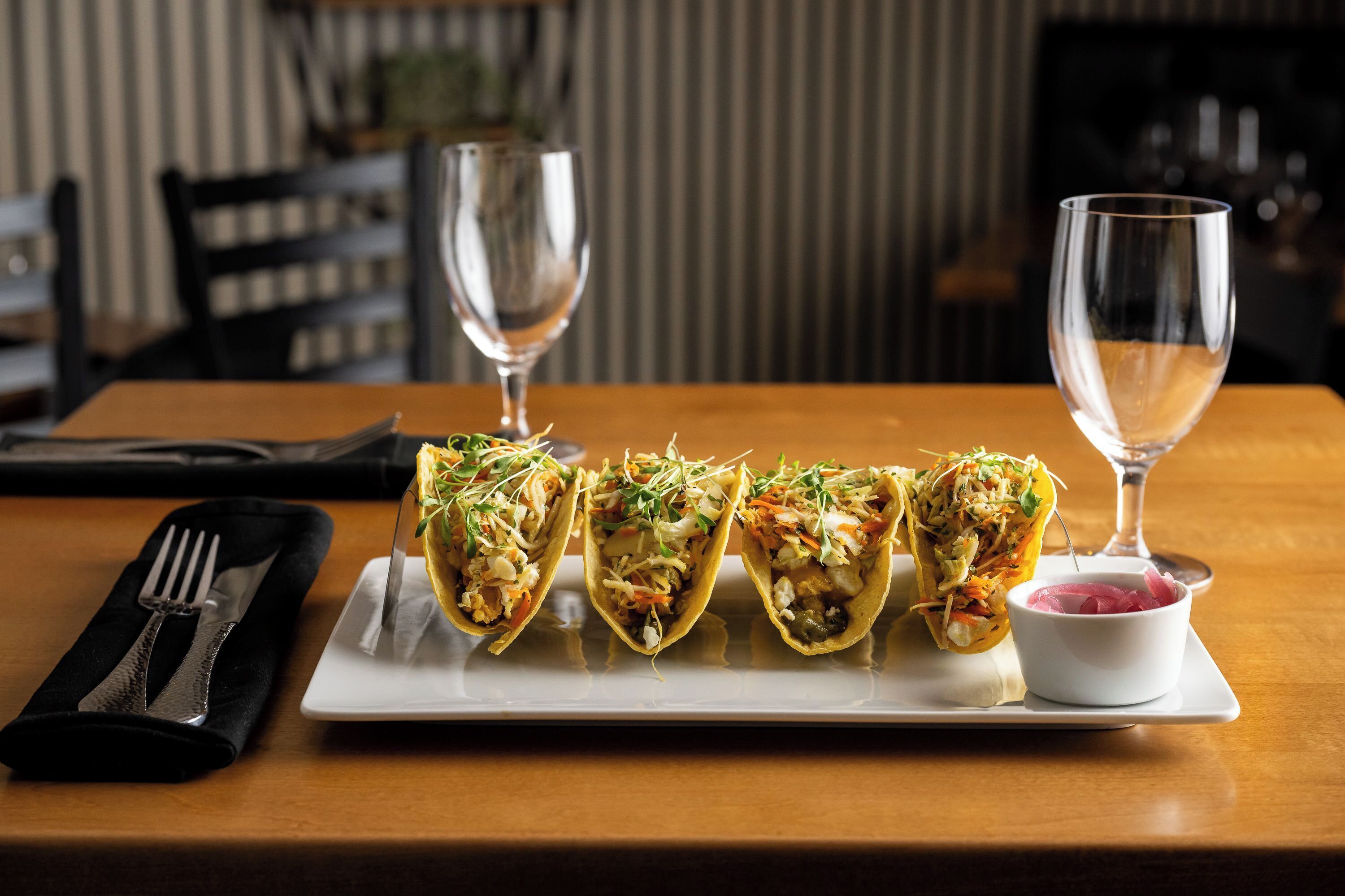Een houten tafel met een wit bord waarop drie taco's liggen en een klein schaaltje ingemaakte uien.