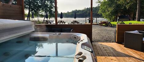 A hot tub with a wooden deck, picnic tables, and umbrellas, overlooking a lake with trees and a cabin in the background.