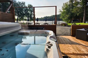 Un jacuzzi con muelle de madera, mesas de picnic y sombrillas, con vistas a un lago con árboles y una cabaña al fondo.