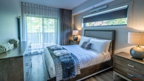 Una habitación de hotel moderna con una cama grande, una mesita de noche, una lámpara y un ventanal con cortinas.