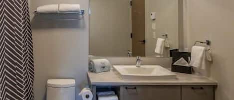 Un baño moderno con inodoro, lavabo y espejo. Hay toallas y artículos de aseo en la encimera.