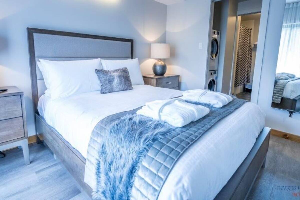 A modern hotel room with a large bed, a bedside table with a lamp, a mirror, and a view of the bathroom.