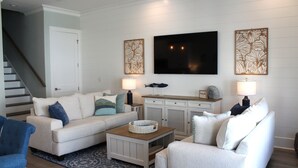 A living room with a sofa, coffee table, and TV. There is a staircase and a cabinet with decorative items.