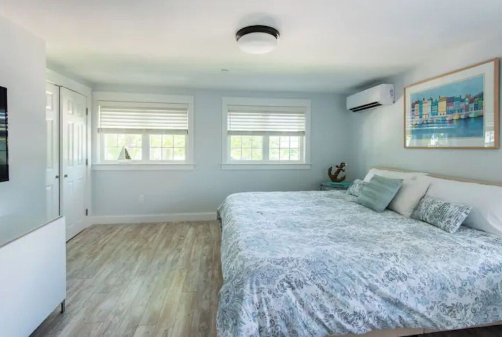 Un dormitorio con una cama, dos ventanas, un equipo de aire acondicionado y un cuadro en la pared.