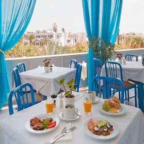 A dining area with tables and chairs, a view of buildings, and a table set with food and drinks. - Hotel Glaros (Santorini)