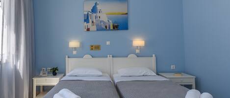Quarto de hotel com duas camas, pintura de uma paisagem costeira, abajures, uma mesinha com um telefone e uma planta, e uma janela com cortinas.