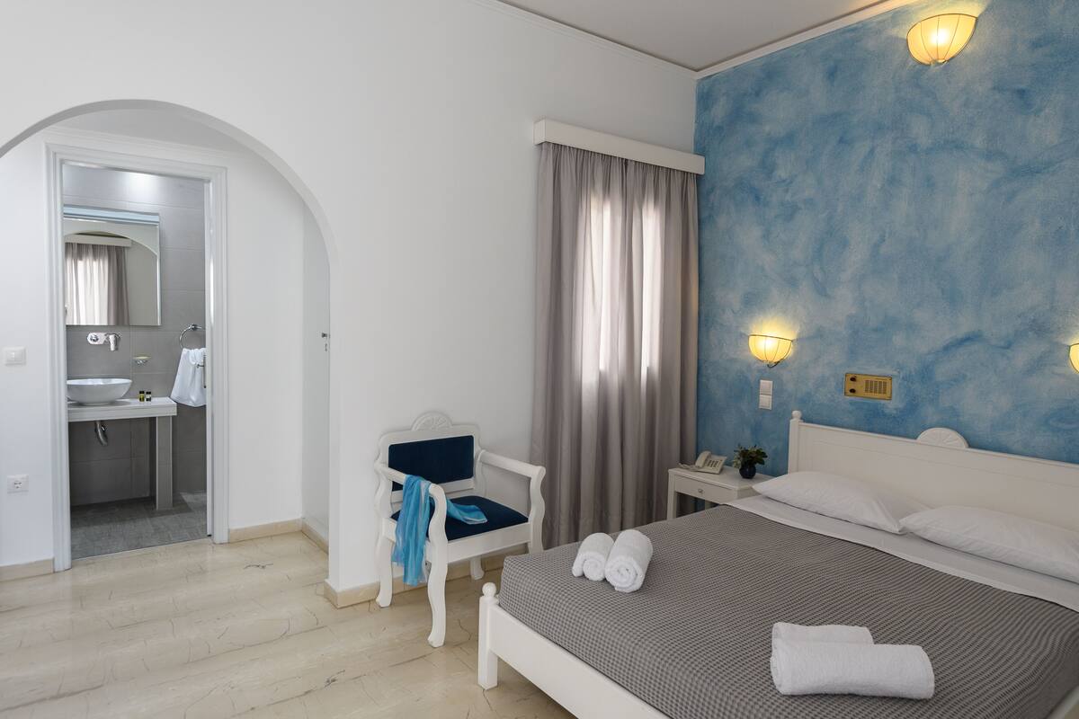 A bedroom with a bed, a nightstand, a chair, a bathroom visible through an archway, and a blue wall.