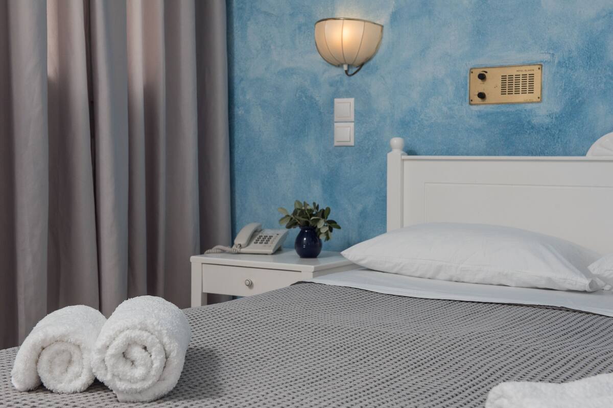 A neatly made bed with white linens, a bedside table with a lamp, a phone, and a vase with a plant.