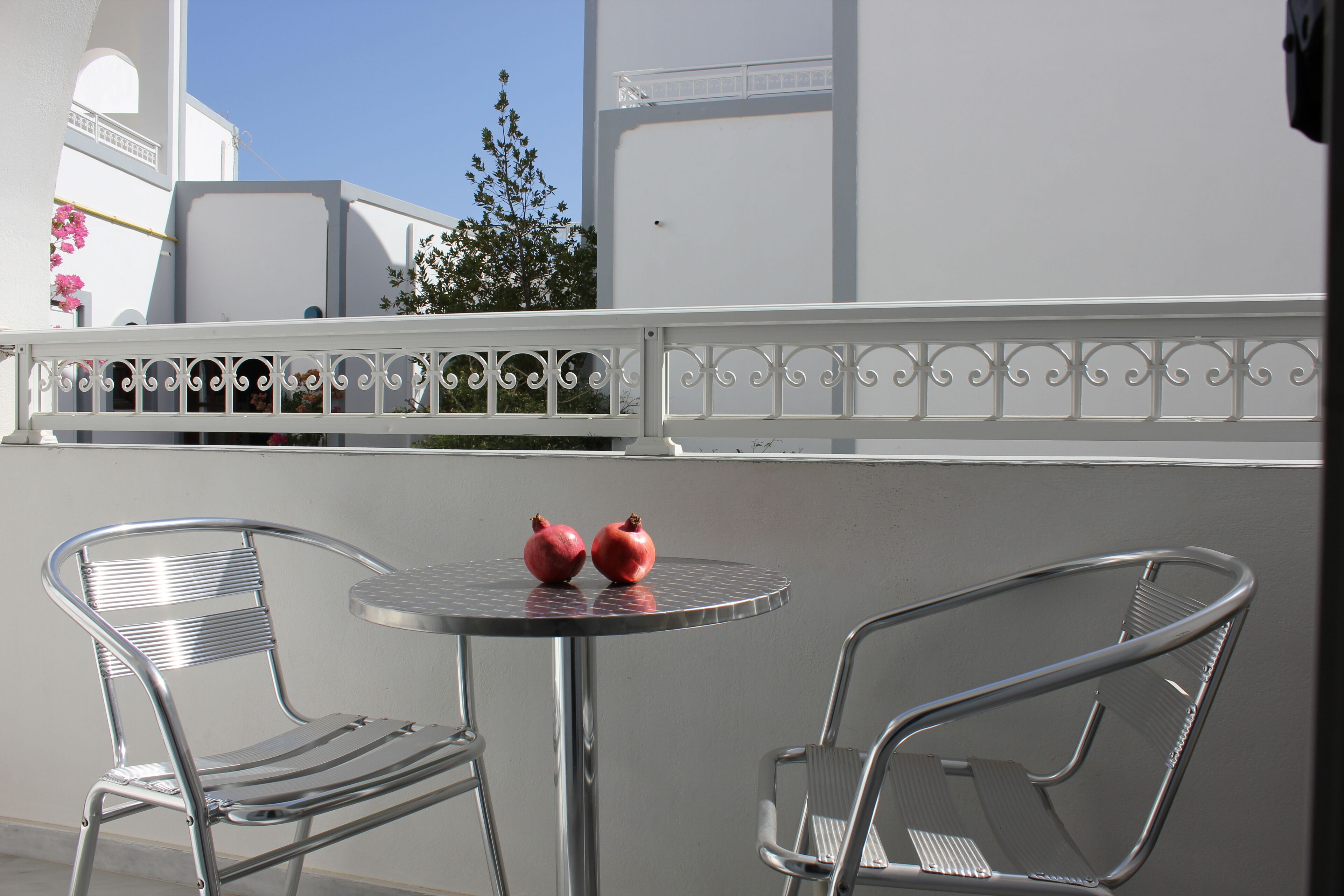 A balcony with two metal chairs and a small round table, two pomegranates on the table, and a white railing with decorative elements.