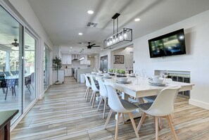 A modern kitchen with a dining area, large windows, and a flat-screen TV.