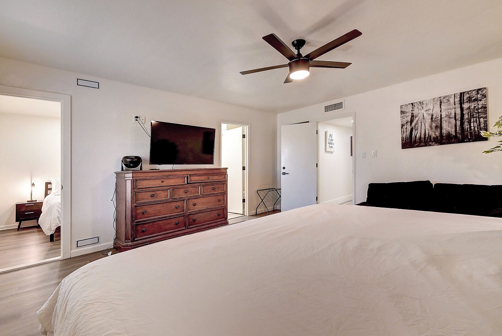 A bedroom with a large bed, a ceiling fan, a dresser, a television, and a painting on the wall.