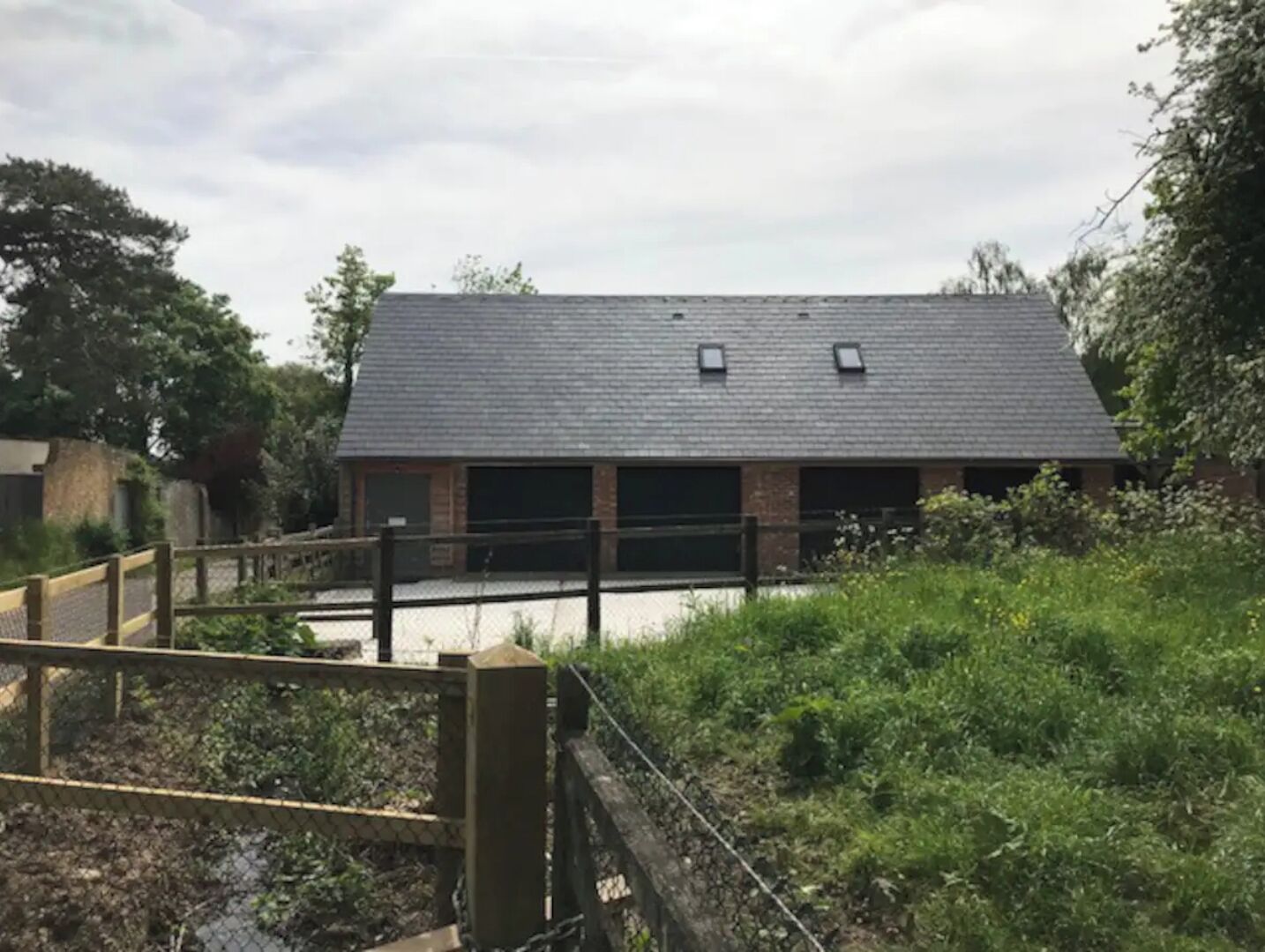 A building with a dark roof and brick walls, surrounded by greenery and a wooden fence.