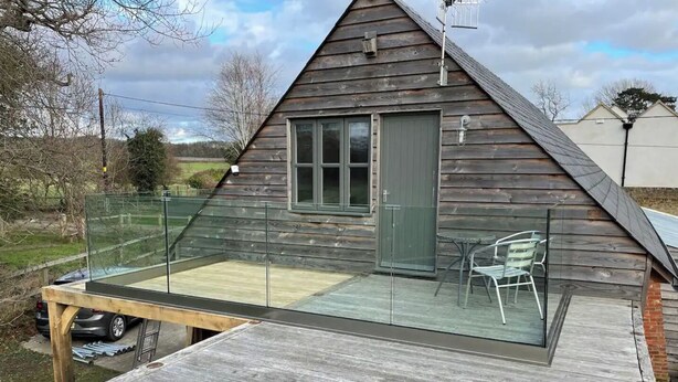Een houten chalet met een glazen balkon, een deur en een kleine tafel met stoelen.