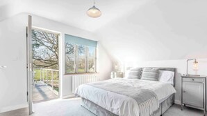 Een slaapkamer met een groot bed, een nachtkastje met een lamp, een raam met uitzicht op bomen en een deur naar een balkon.