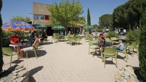 An outdoor café with tables and chairs, surrounded by trees and a building with a red door. - La Roque d'Anthéron - Mobil-home - 6 pers - 3 bdrms - Swimming pool (La Roque-d'Anthéron)