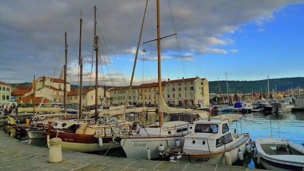 Una marina con diverse imbarcazioni ormeggiate, edifici sullo sfondo e cielo sereno.