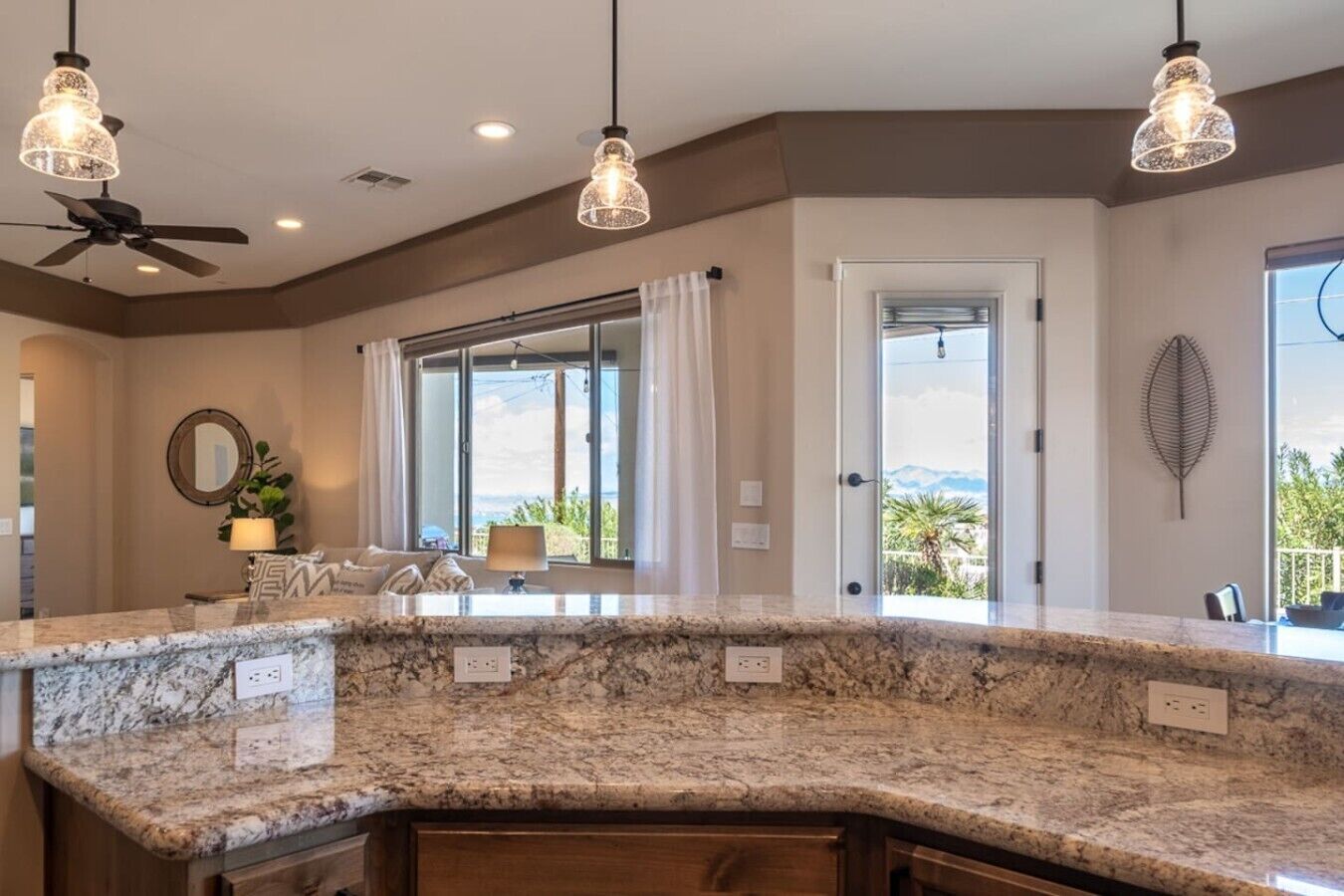 A kitchen with a granite countertop, pendant lighting, a ceiling fan, and a view through a large window.
