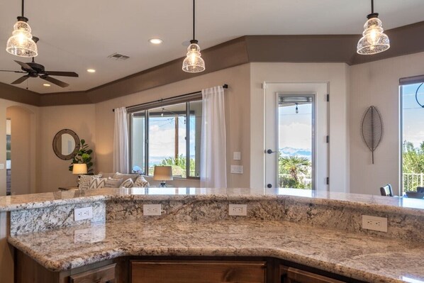 A kitchen with a granite countertop, pendant lighting, a ceiling fan, and a view through a large window.