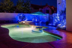 A swimming pool with a curved design, surrounded by a patio area with a blue-lit fence and a single umbrella. - Lake View Oasis Pool/Spa+Boat Garage+Ice Machine (Lake Havasu City)