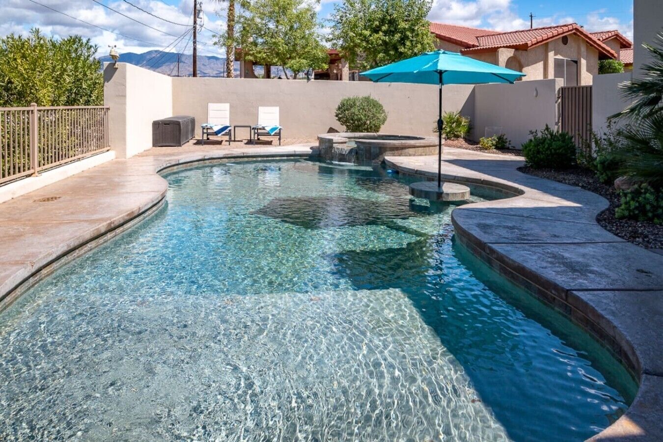 A clear swimming pool with a stone edge, a blue umbrella, and lounge chairs.