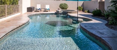 A clear swimming pool with a stone edge, a blue umbrella, and lounge chairs.