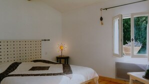 Un dormitorio con cama, una mesita, una ventana con vistas a zonas verdes y una lámpara fijada a la pared.