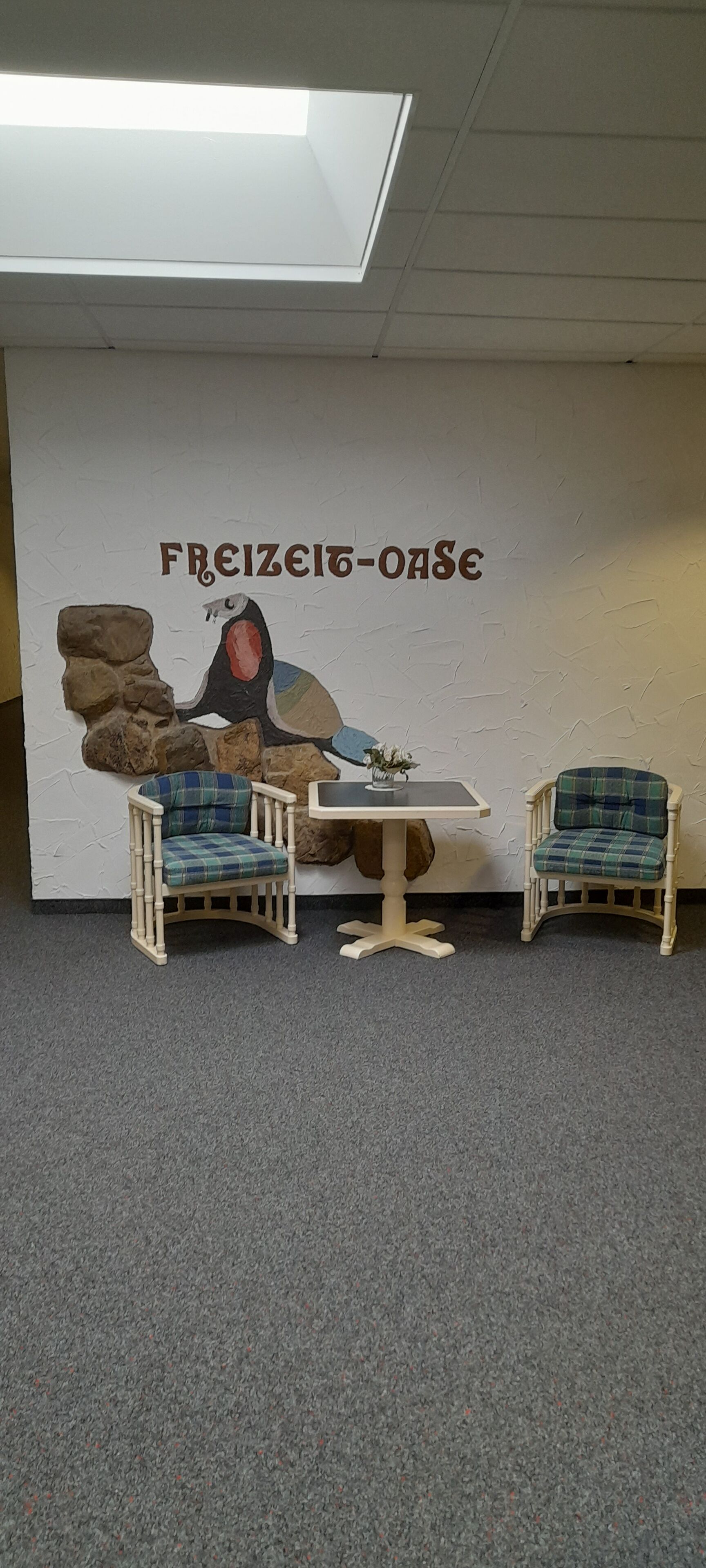 A room with two wooden chairs with plaid cushions, a small table, and a wall with a mural and the text "FREIZEIT-OASE".