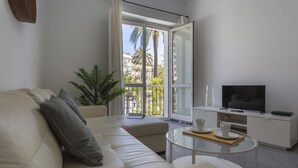 A modern living room with a sofa, a glass coffee table, a TV, and a balcony with a view of palm trees and buildings.