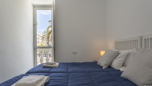 A bedroom with a large bed, a window with a view of palm trees, and a bedside table with a lamp.