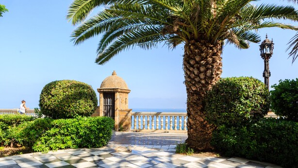 A checkered pathway leading to a building with a dome, surrounded by greenery and a view of the sea.