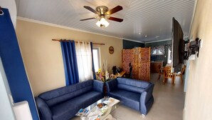 A living room with a blue sofa, a coffee table with magazines, a ceiling fan, and a view into the kitchen.