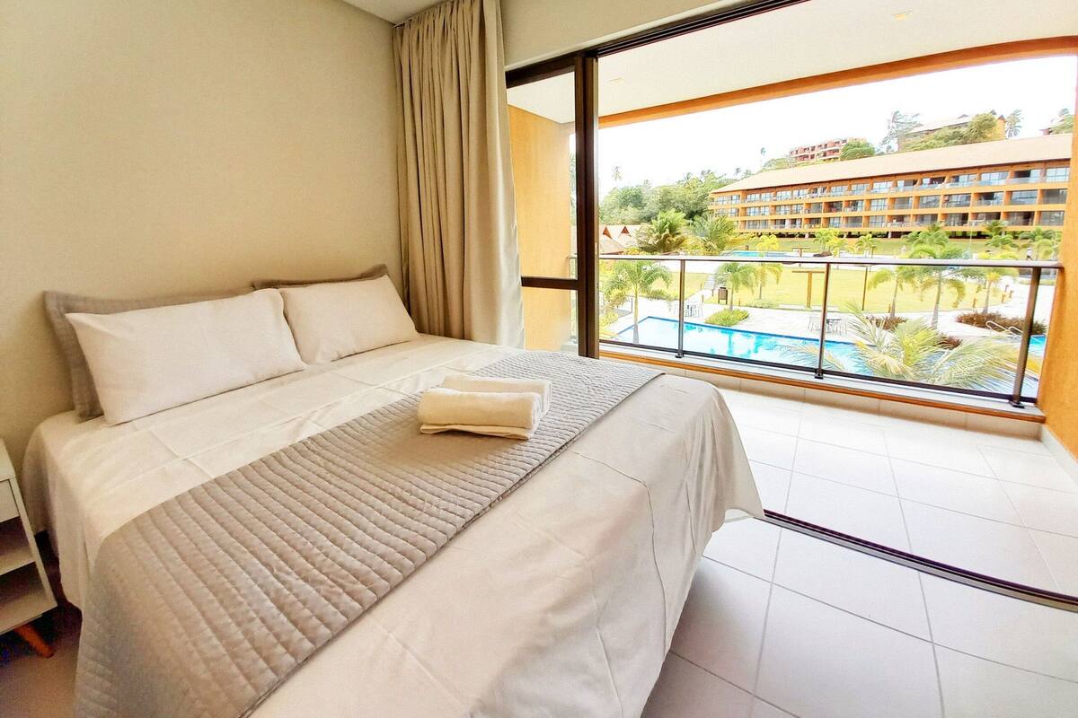 A hotel room with a large bed, a view of a pool and buildings, and a balcony with curtains.