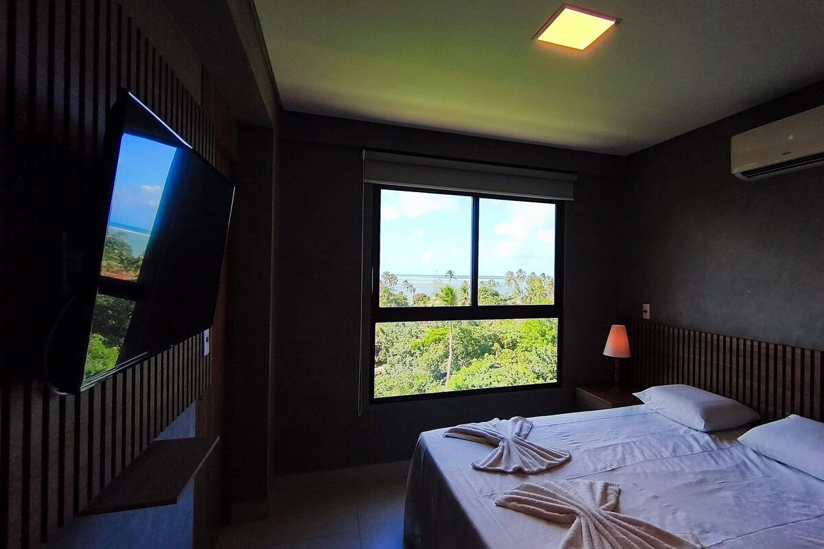A hotel room with a bed, a TV, a window with a view, and an air conditioning unit.