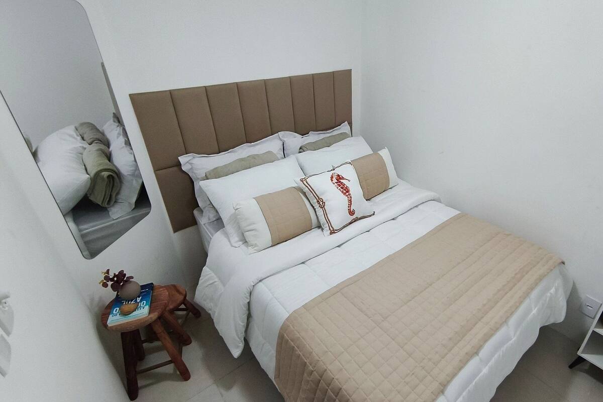 A neatly made bed with a headboard, a small wooden stool with a book, and a shelf with decorative items.