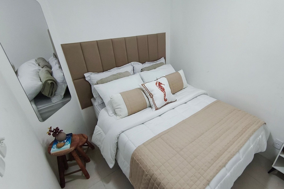 Cama bem arrumada com cabeceira, um banquinho de madeira com um livro e uma prateleira com objetos decorativos.