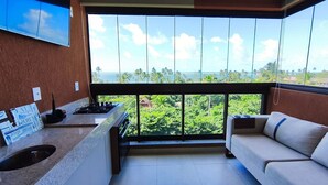 A balcony with a view of the ocean and palm trees, a small kitchen area with a gas stove, and a sofa.