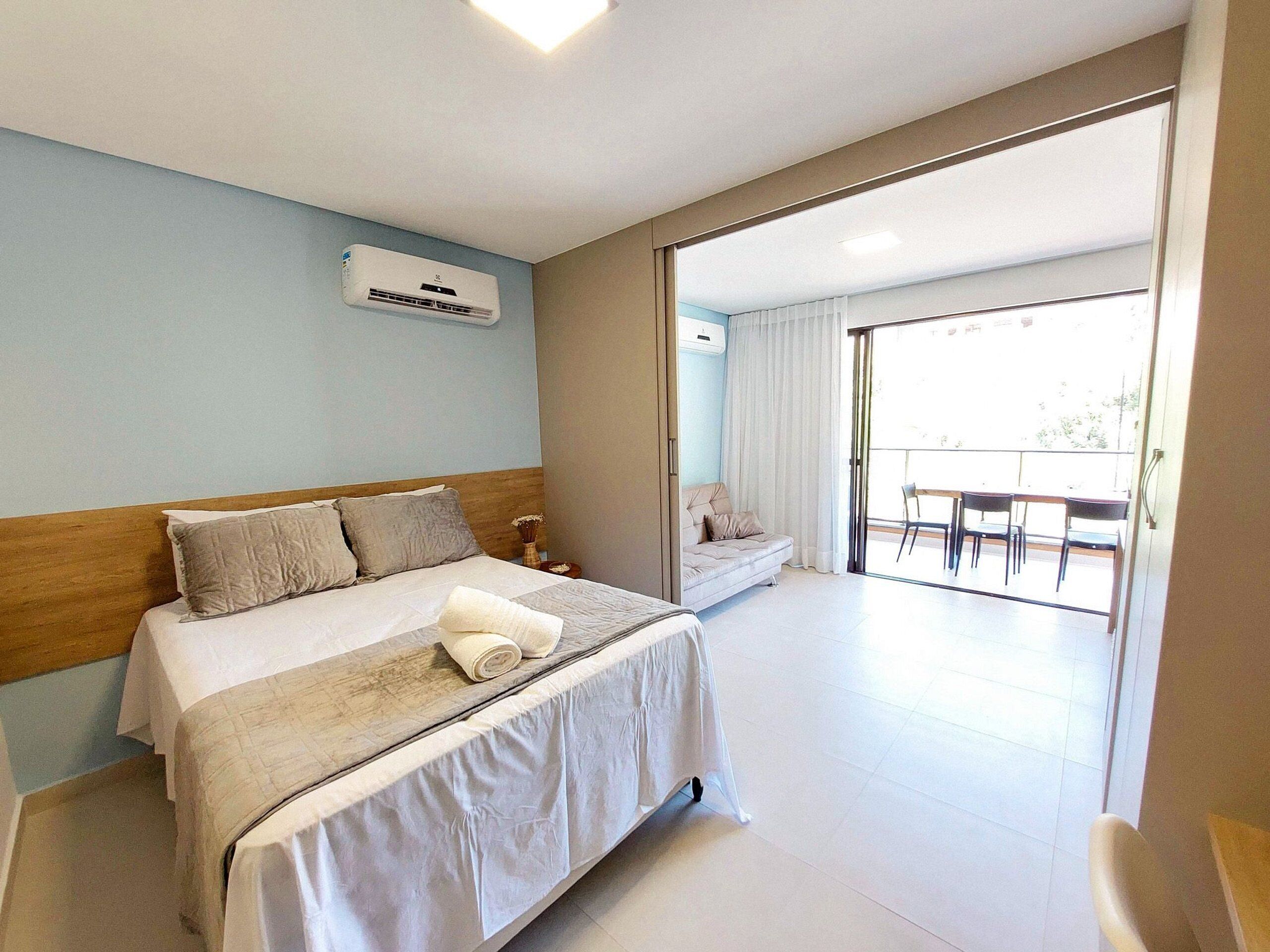 Habitación de hotel con cama, mesita de noche, silla, balcón con mesa y sillas, y un equipo de aire acondicionado en la pared.
