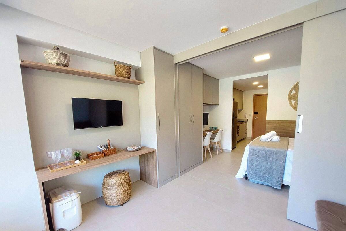 A modern hotel room with a flat-screen TV, a bed, a desk with a chair, and a kitchenette.