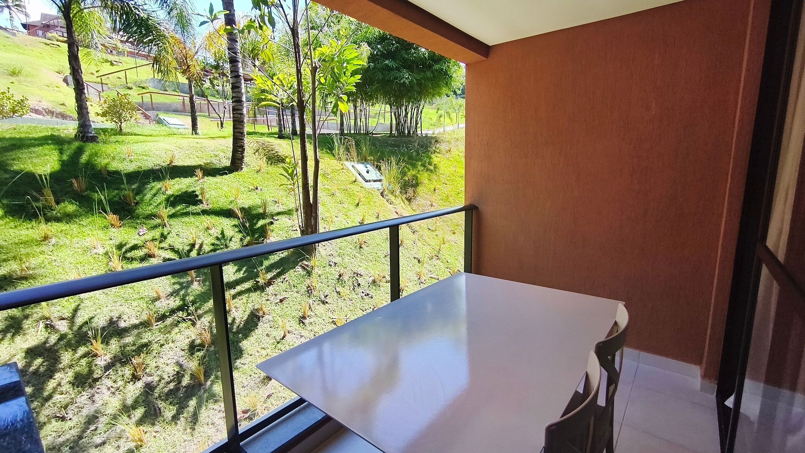 Un balcón con barandilla de metal, una mesa blanca y vistas a una zona verde con árboles.