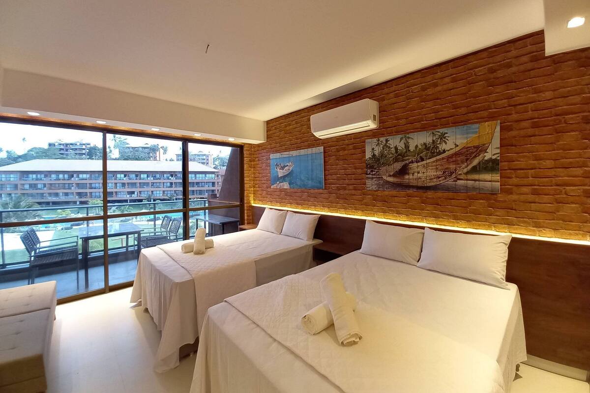 A hotel room with two beds, a large window with a view of buildings, a brick wall, and artwork.