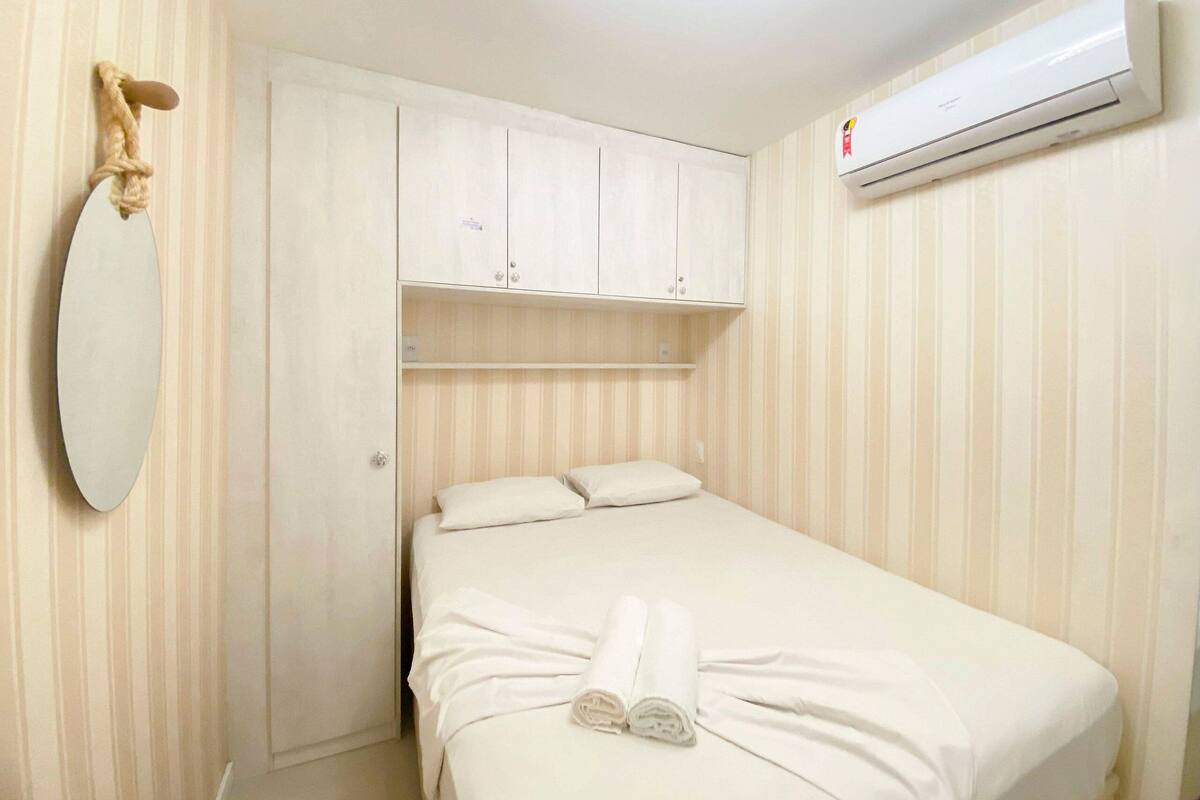 A small, single-bed room with a white bed, a mirror, a wardrobe, and an air conditioning unit.