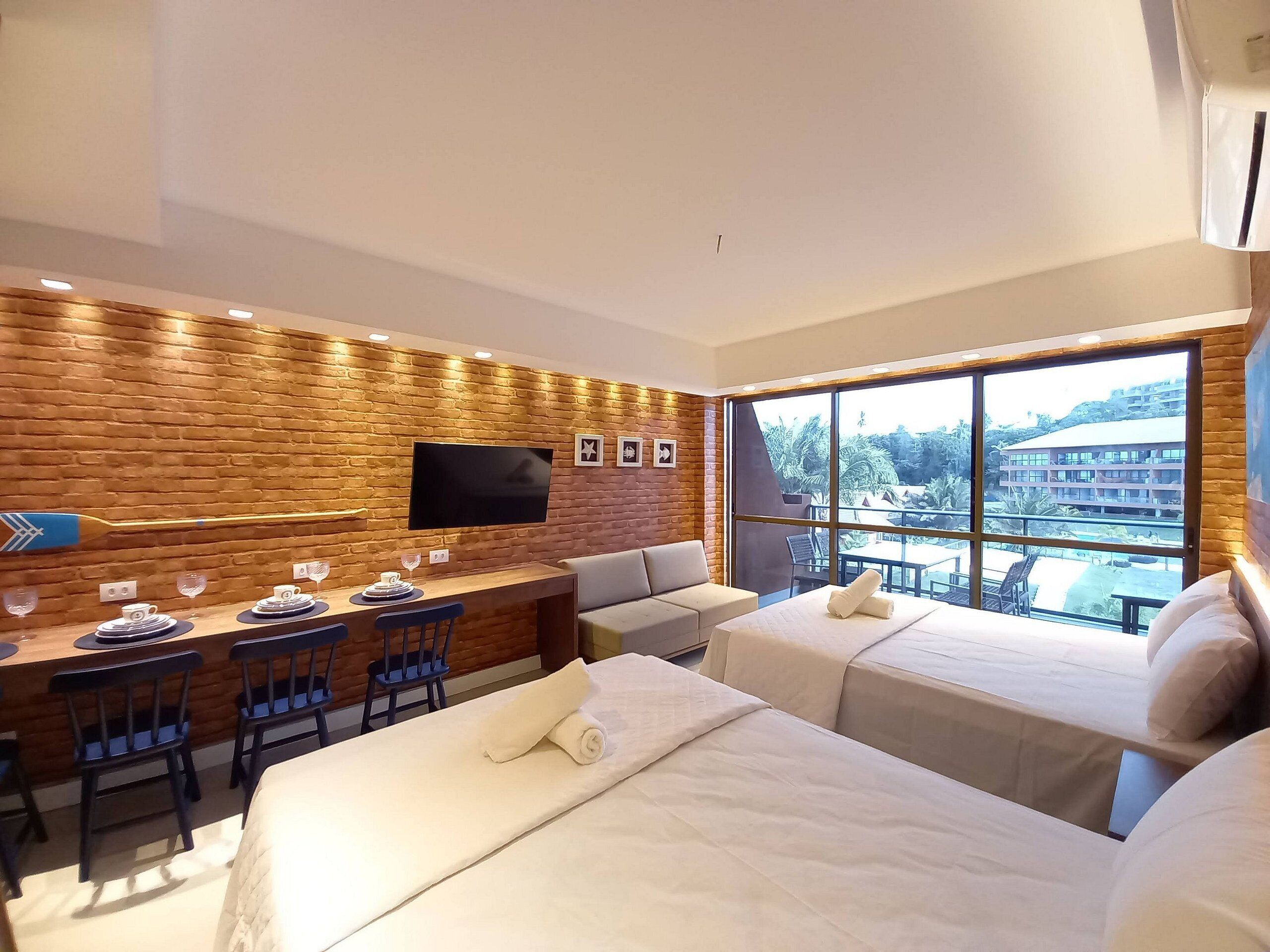 A hotel room with a brick wall, a flat-screen TV, a dining area with chairs and a table, two beds, and a large window with a view of buildings and greenery.