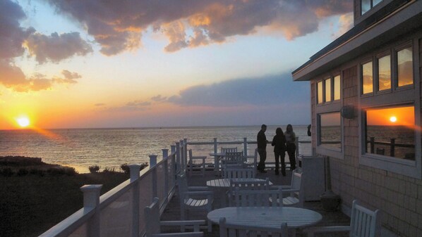 A seaside terrace with tables and chairs, overlooking the ocean at sunset.