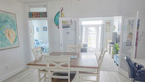 Un coin repas avec une table en bois et des chaises blanches, un ventilateur de plafond et une vue sur une chambre à coucher avec un lit et des rideaux.